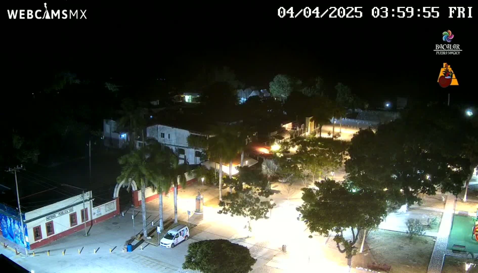 A nighttime aerial view of a town square with palm trees and buildings illuminated by streetlights under a clear sky.