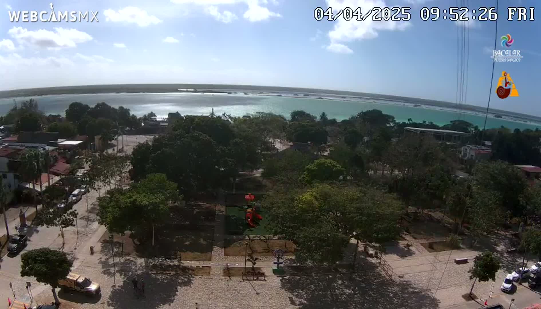 An aerial view of a town square with lush green trees, a playground, and parked cars, overlooking a calm, turquoise body of water under a partly sunny sky.