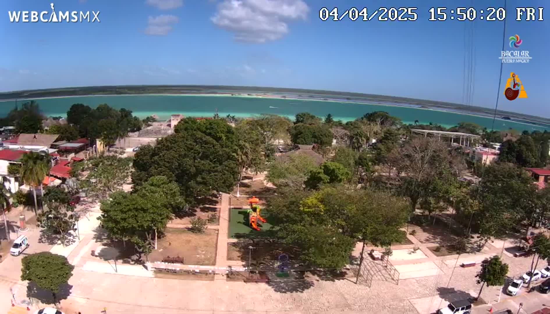 An aerial view shows a town square with a playground, surrounded by lush green trees, next to a calm, turquoise body of water under a sunny sky.