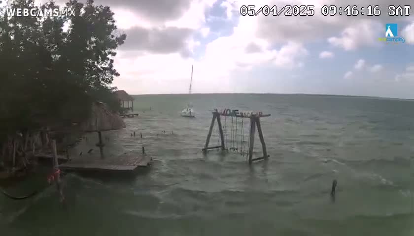 A partly cloudy day shows a choppy body of water with a small sailboat, a partially submerged swing set, and a dock near a shoreline with trees and a structure.