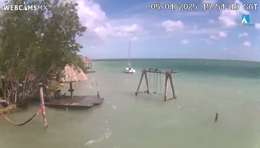 A partly cloudy day shows a tranquil body of light green water with a sailboat, a set of swings in the water, and a shoreline with trees and a small dock under a thatched roof.