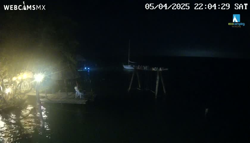 A nighttime view of a waterfront scene shows a sailboat moored near a dock with lights illuminating a partially visible structure and surrounding water.
