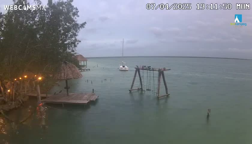 A tranquil body of water under a cloudy sky features a sailboat, a wooden swing set, and a small dock with a thatched umbrella near a shoreline with lush vegetation.