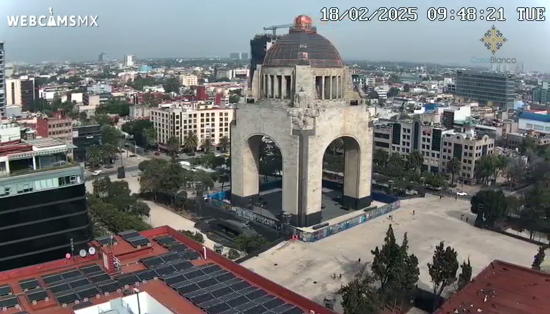 A large stone monument with an archway stands in a city square on a partly cloudy day.
