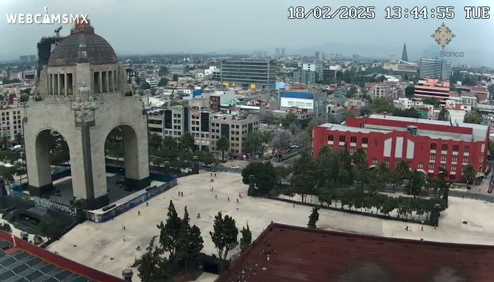 An elevated view shows a large stone monument in a city square on an overcast day, with numerous buildings and trees surrounding it.