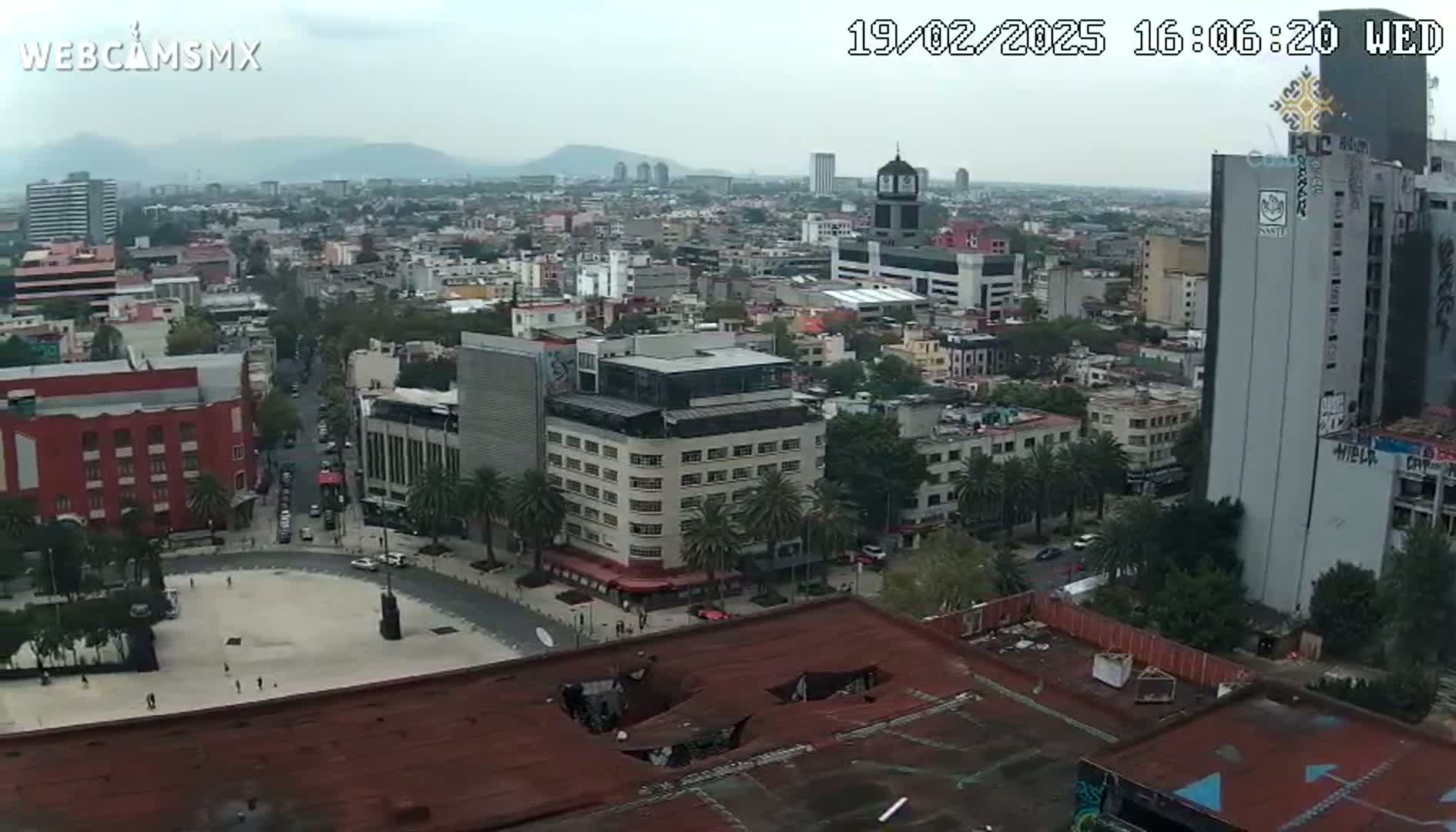 A high-angle, long shot of a city under an overcast sky shows a large plaza and numerous buildings, including high-rises and smaller structures.