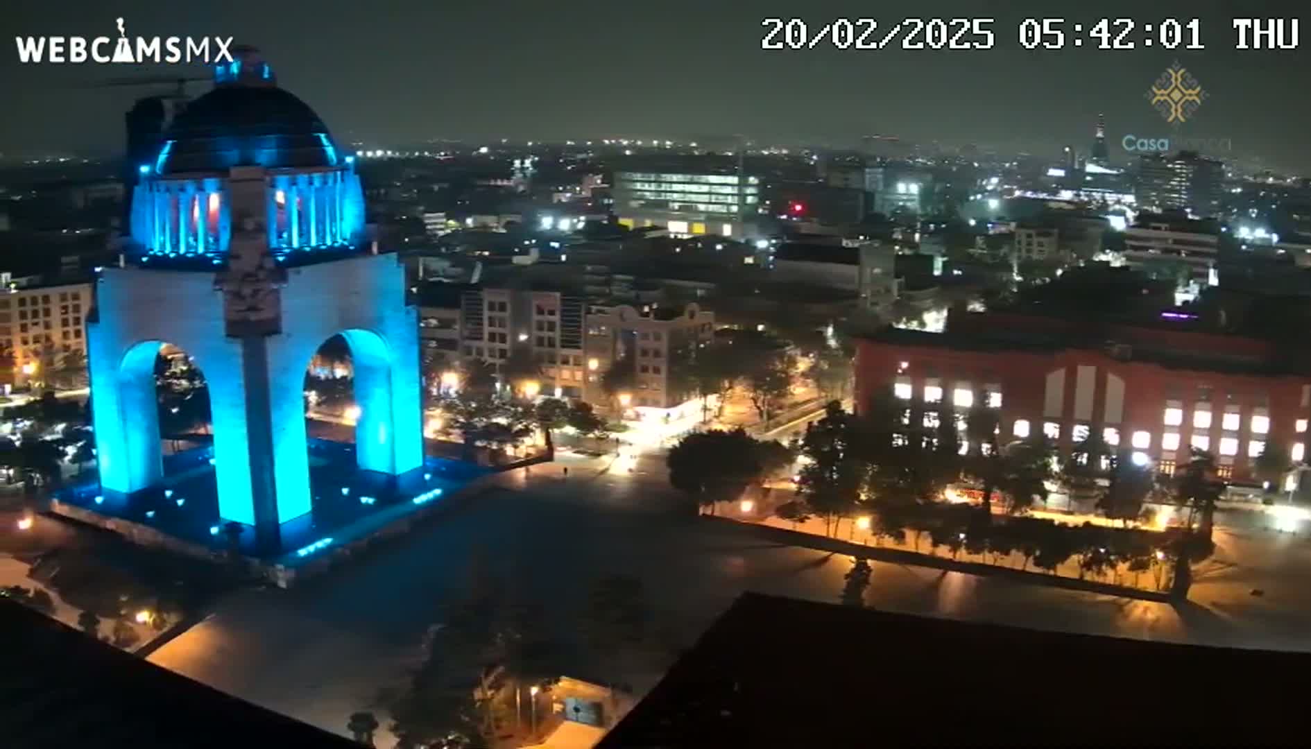 A brightly lit, blue monument stands in a city square at night, surrounded by illuminated buildings and trees under a dark sky.