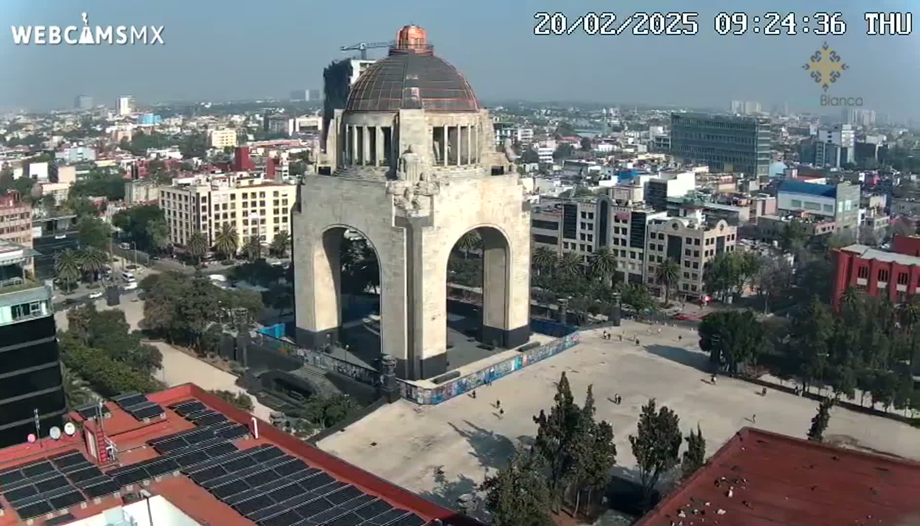 A large stone monument with an archway stands in a city square on a sunny day, surrounded by buildings and trees.