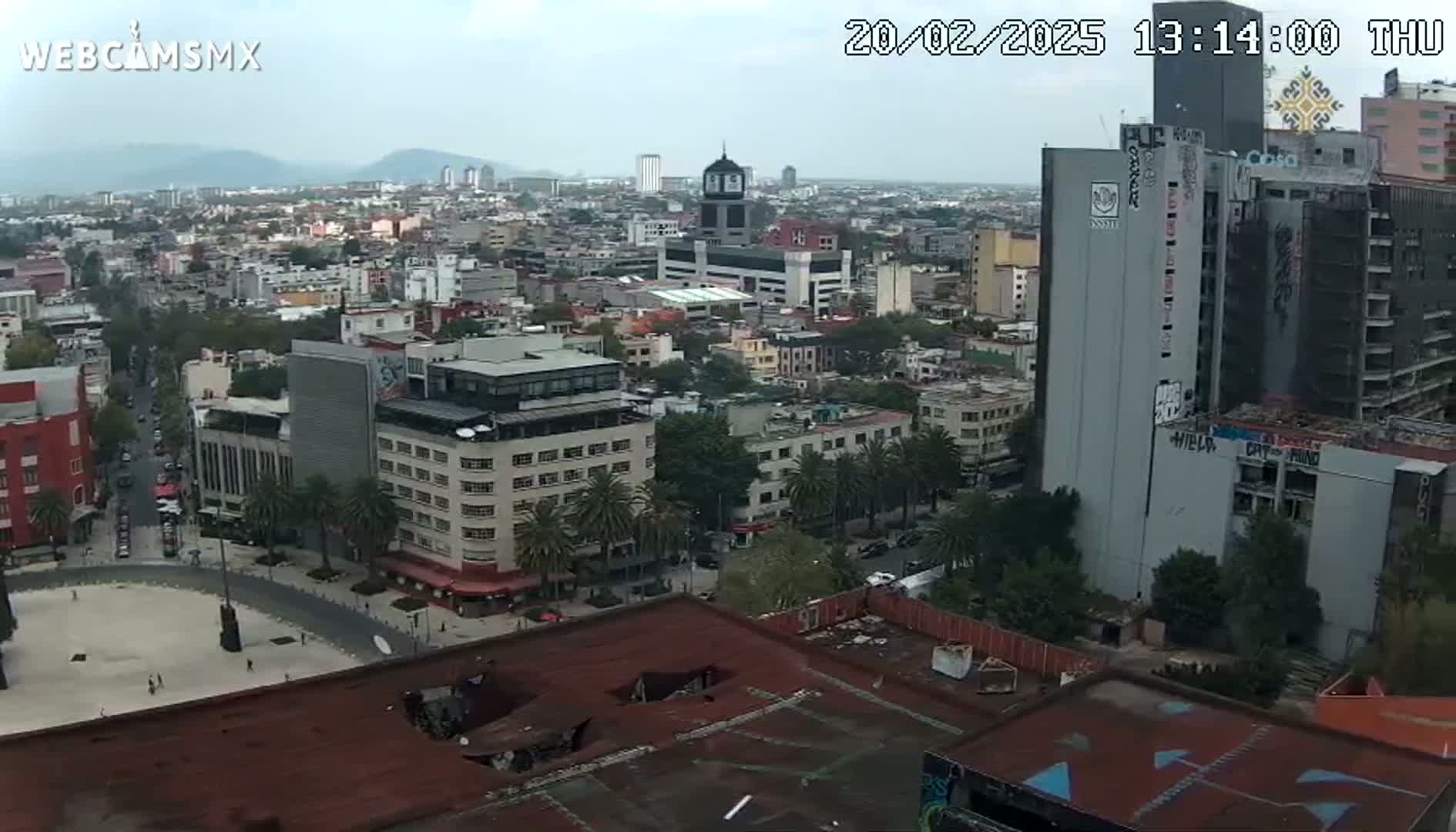 A high-angle, long shot of a city on an overcast day shows a cityscape with several buildings, a plaza, and a large square in the foreground with trees, palm trees and streets.