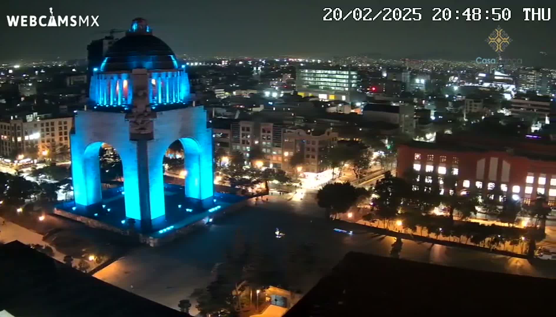 A large monument, illuminated in bright blue, stands prominently in a nighttime cityscape under a clear sky.