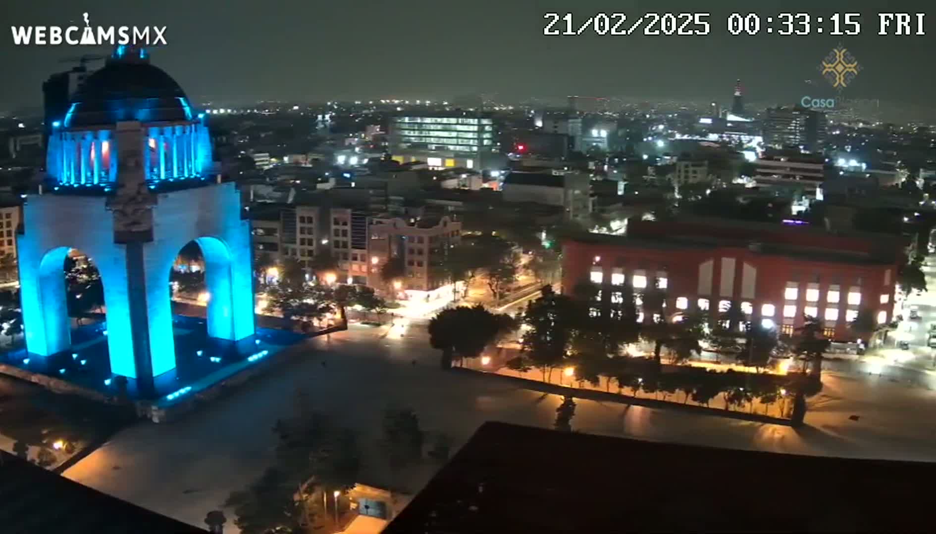 A nighttime, high-angle view shows a monument illuminated in blue light, surrounded by a city with buildings and streets showing ambient light.