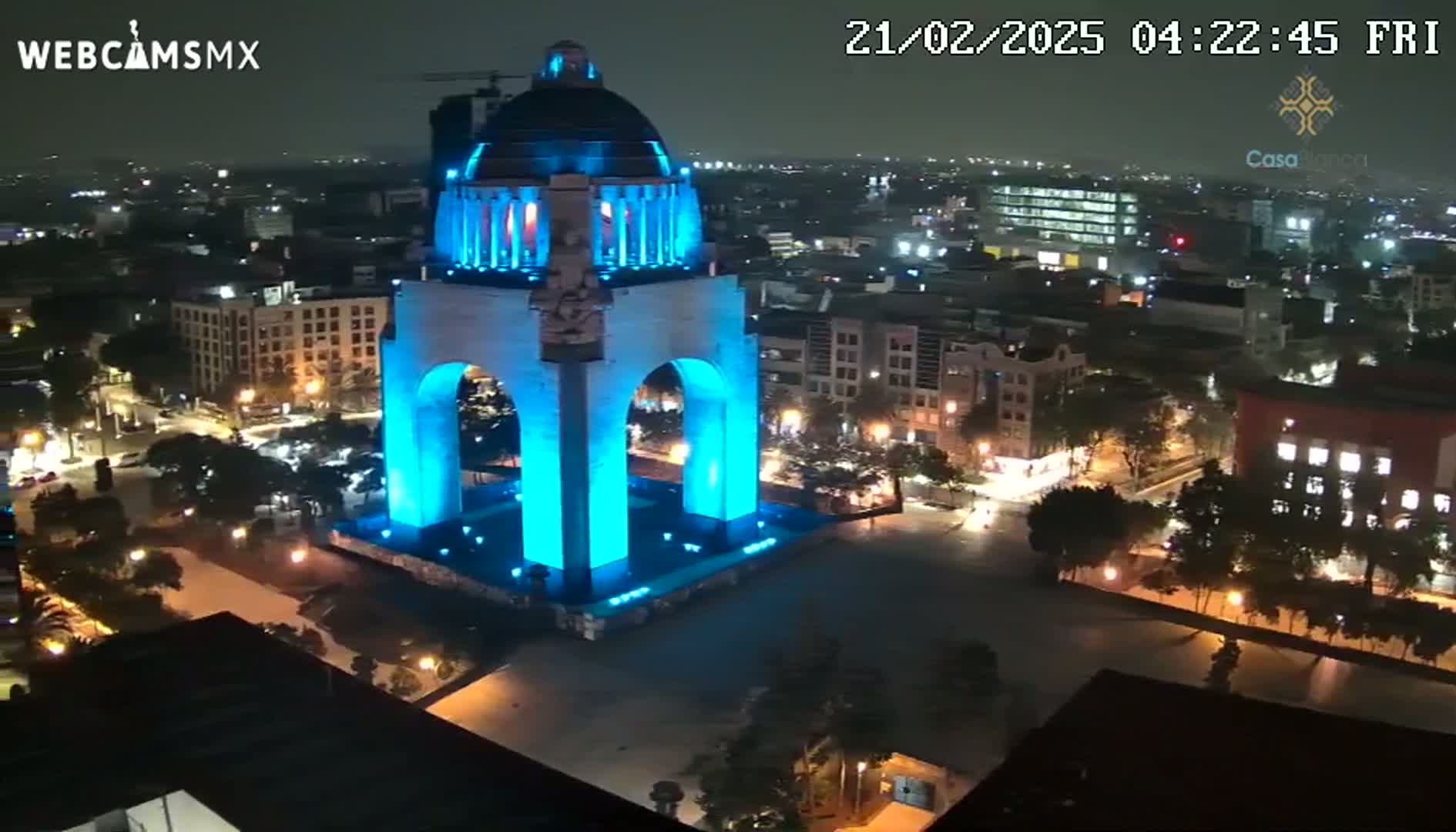 A large monument is illuminated in bright blue at night, surrounded by a city with streetlights visible.