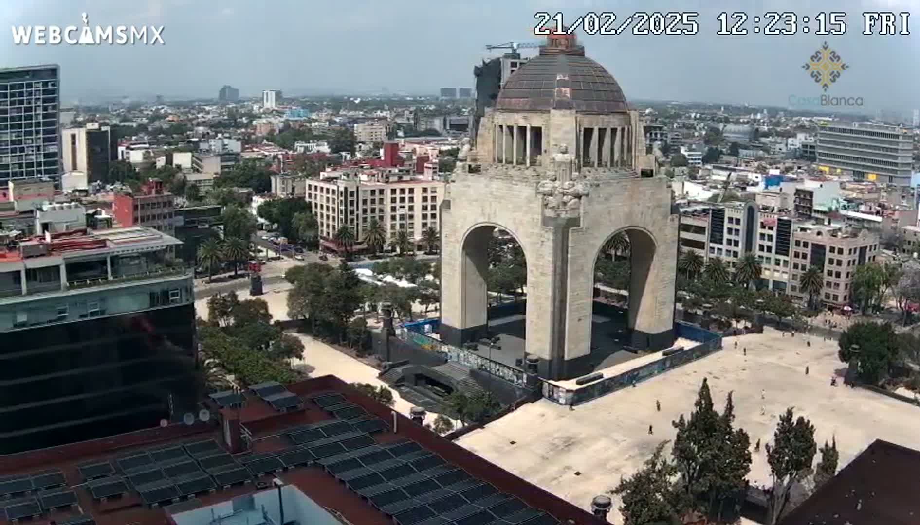 A large stone monument with an archway stands in a mostly empty plaza, surrounded by a city under a partly sunny sky.