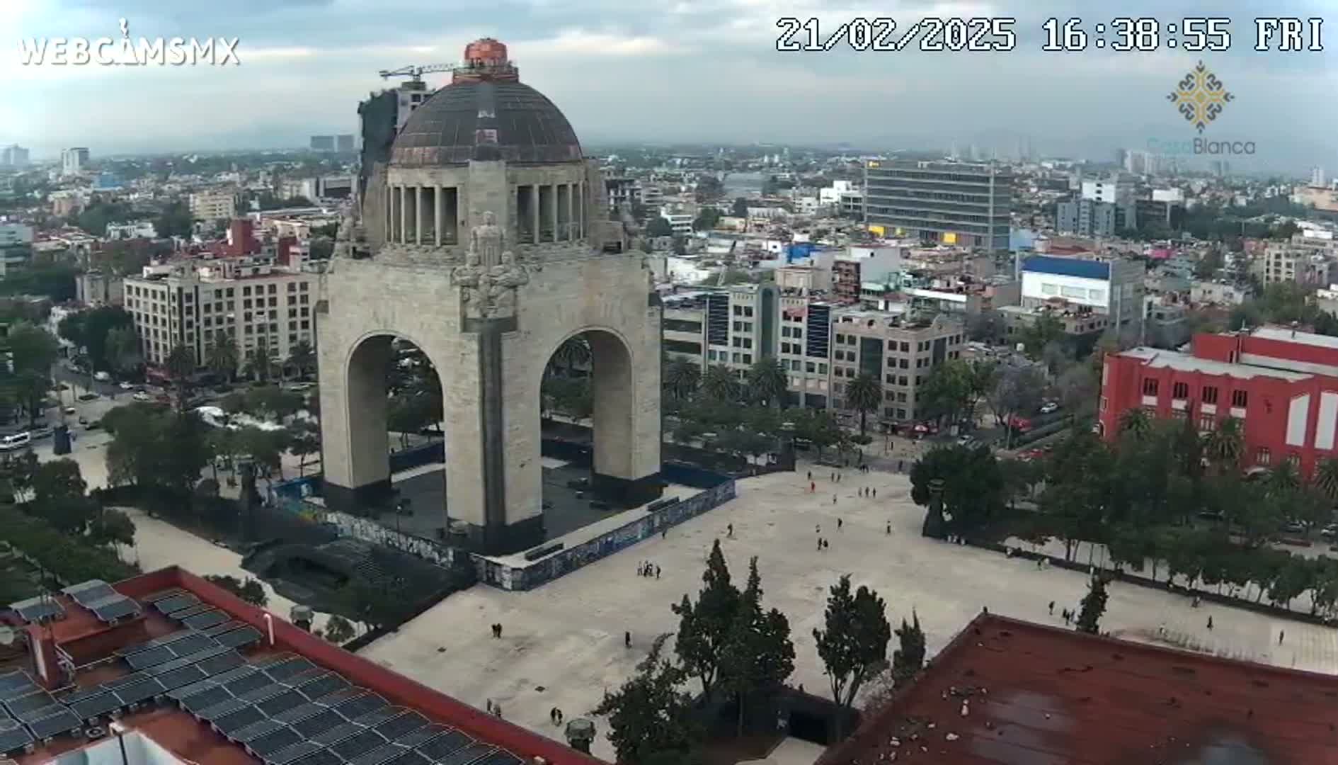 A large stone monument with an archway stands in a city square on an overcast day, surrounded by buildings and sparsely populated areas of greenery.