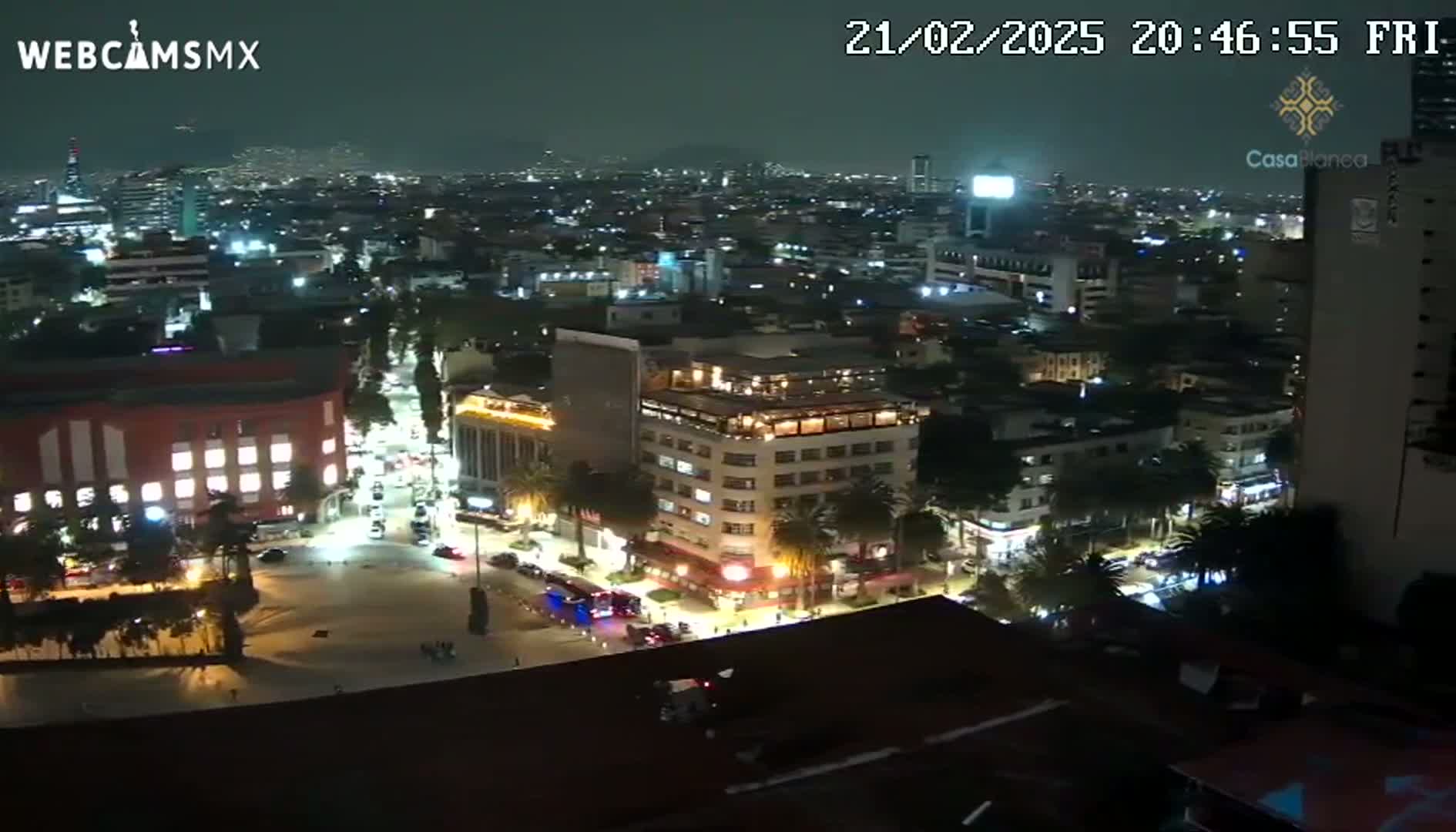 A nighttime, high-angle view of a city with illuminated buildings and streets, showing a large square in the foreground.