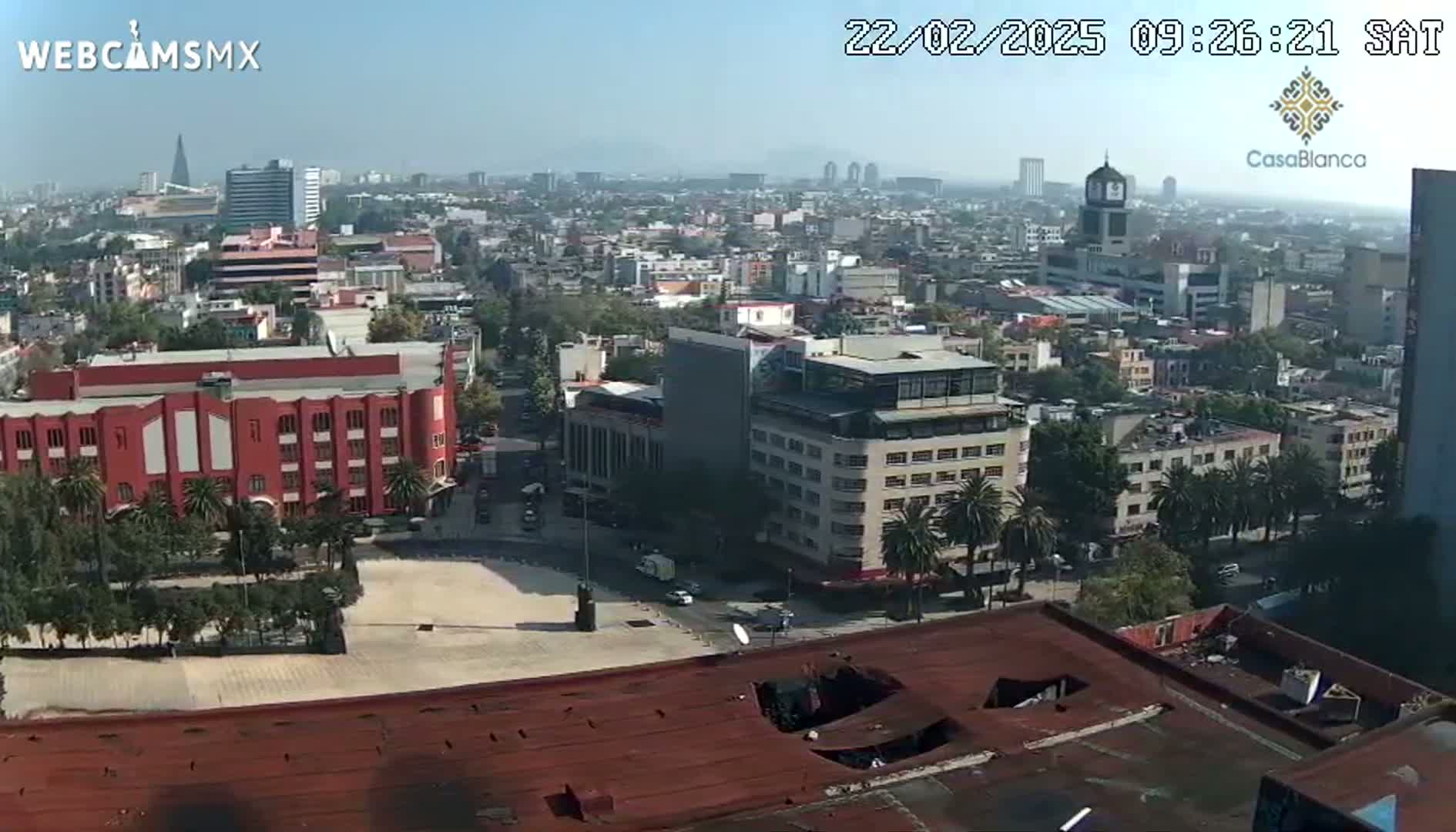 A high-angle, long shot of a city on a hazy day shows a large plaza, red buildings, and a cityscape extending to the horizon.