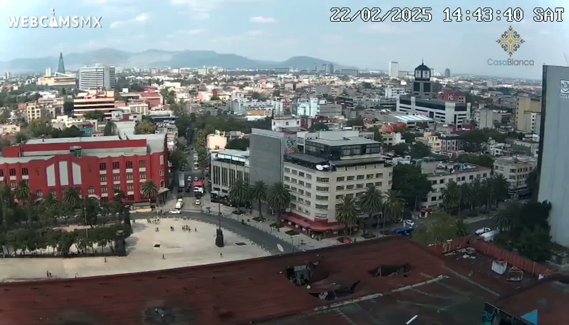 A high-angle, long shot of a city on a partly cloudy day shows numerous buildings, streets, and a large square.