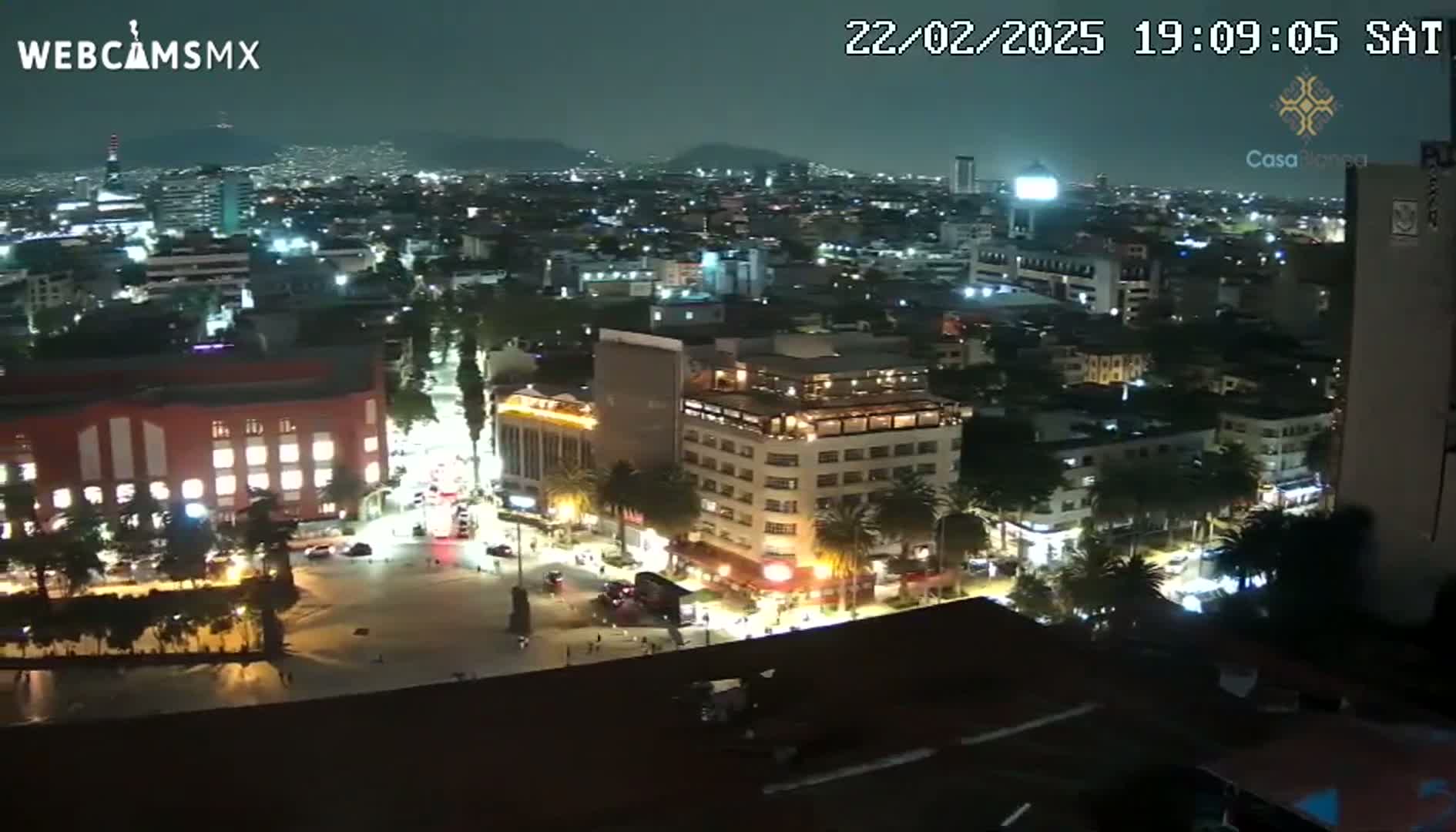 A nighttime, high-angle view of a city square and surrounding buildings, illuminated by streetlights and building lights under a clear sky.