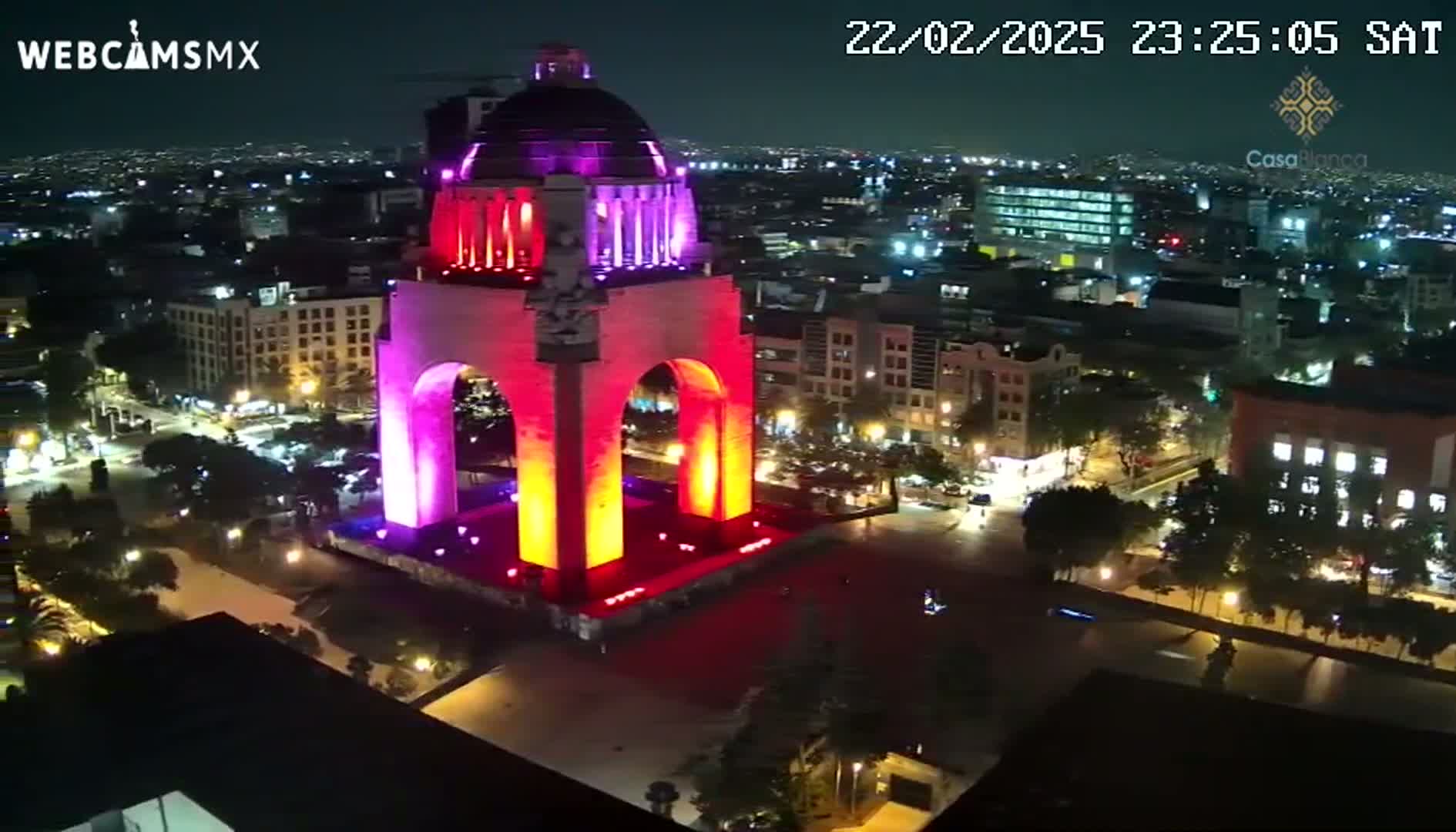 A large monument, illuminated in pink and orange, is centrally located in a nighttime city scene.