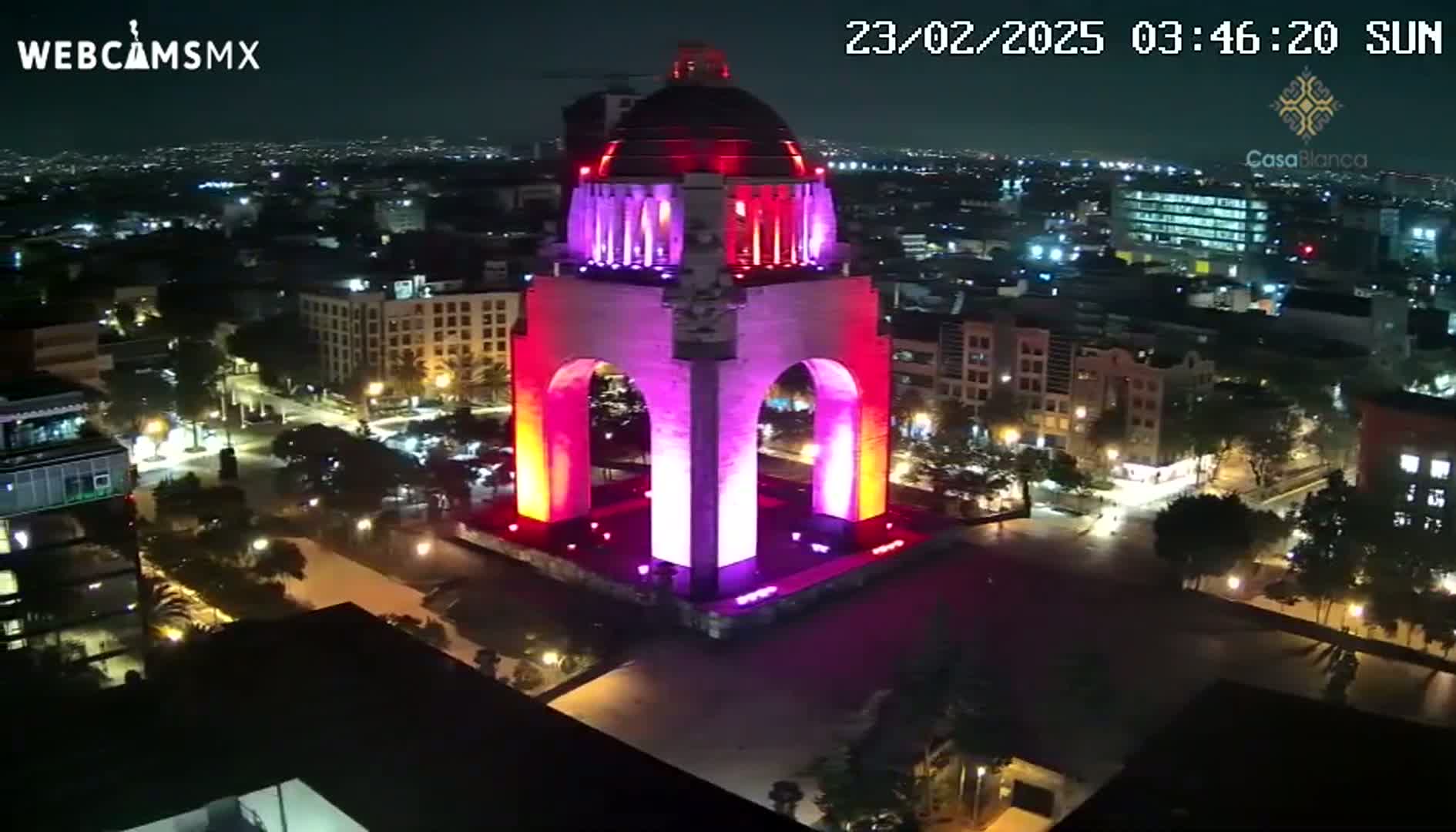 A large monument, illuminated in shades of pink and red, is prominently featured in a nighttime aerial view of a city under a clear sky.