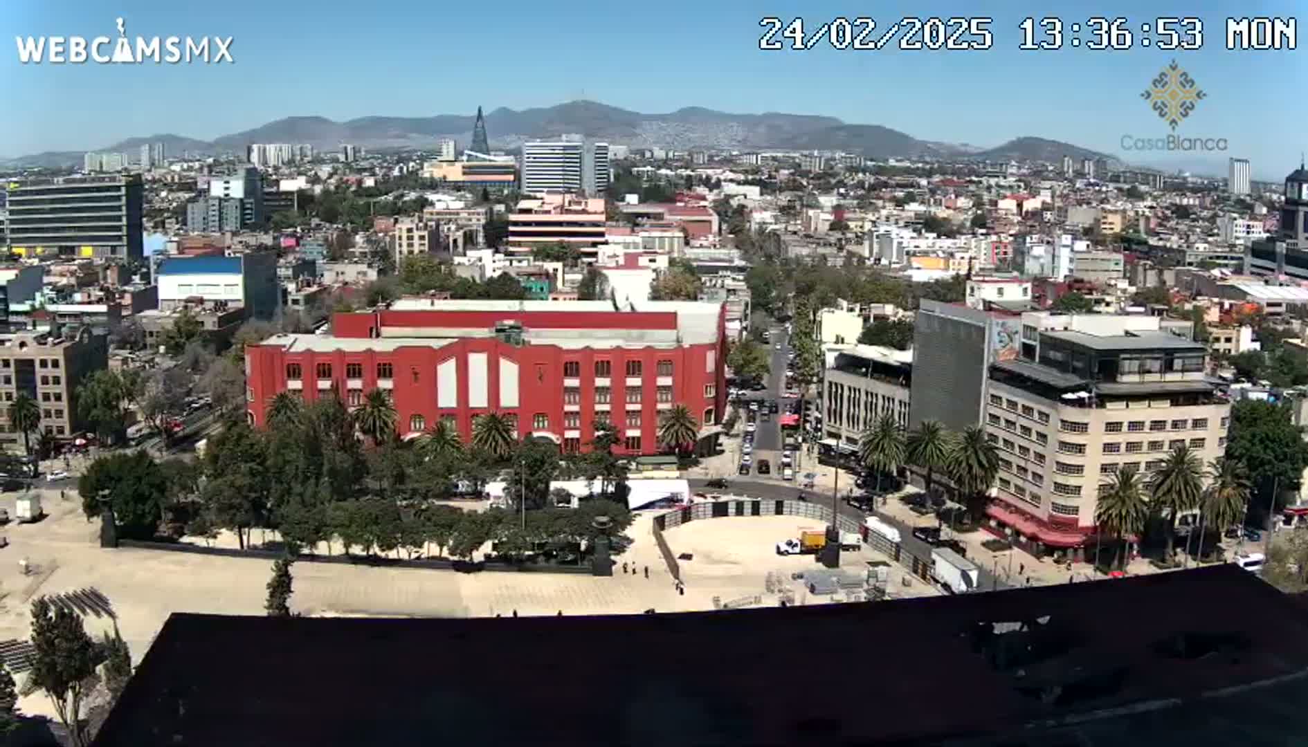A high-angle, long shot of a city on a clear, sunny day shows a large plaza with a red building, surrounded by other buildings and palm trees.