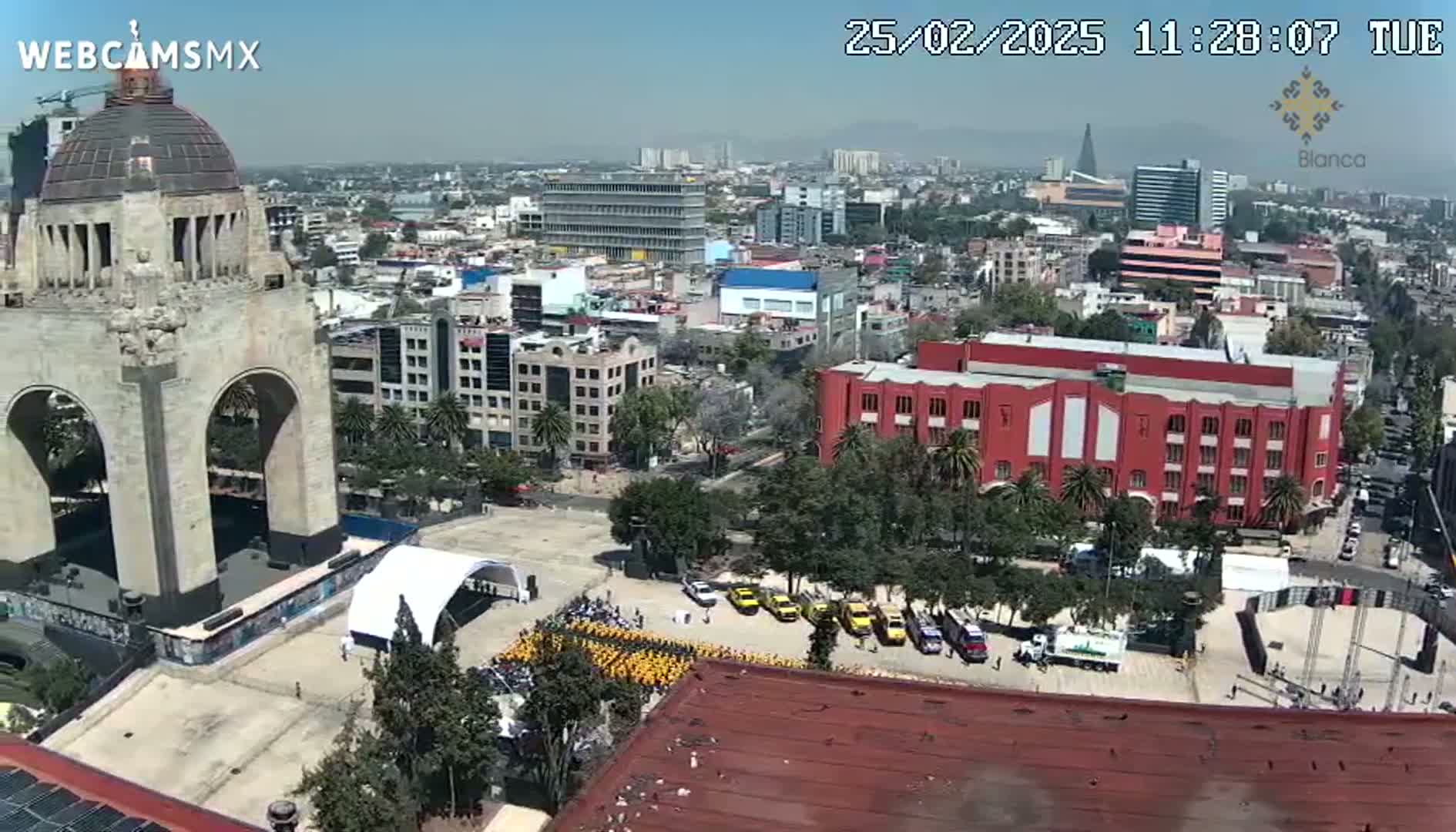 A high-angle, sunny view shows a large city square with a monument, a gathering of people in yellow shirts, and numerous buildings, including a large red building.