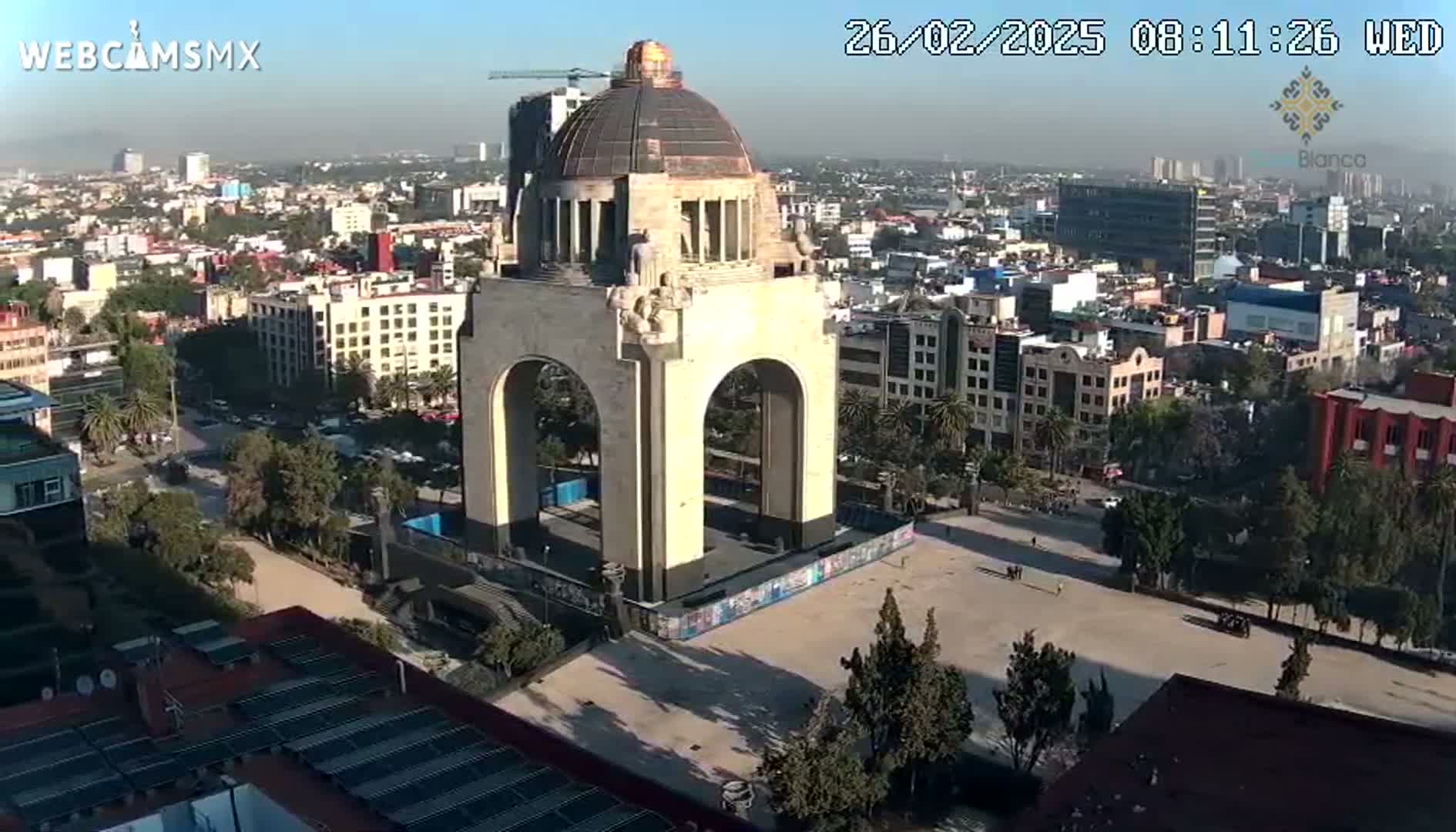 A large monument with a dome and arches stands in a city square on a hazy, sunny day.