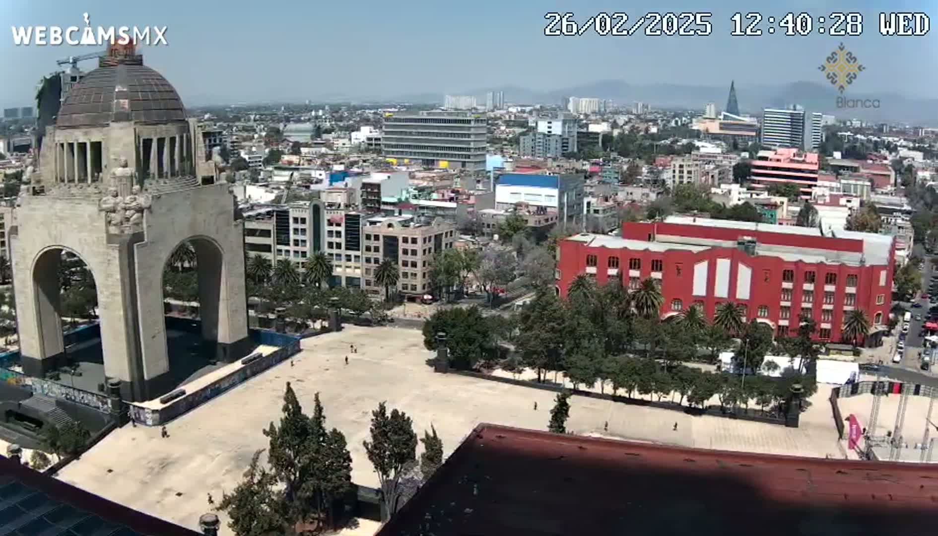 A large stone arch monument stands in a plaza surrounded by a city under a clear, sunny sky.