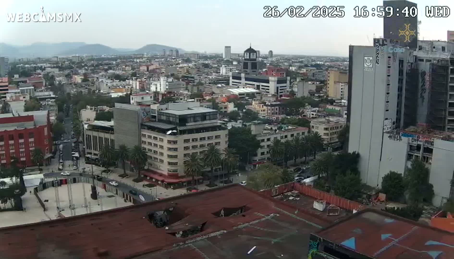 A high-angle, long shot of a city on an overcast day shows a cityscape with many buildings, palm trees, and a large plaza.