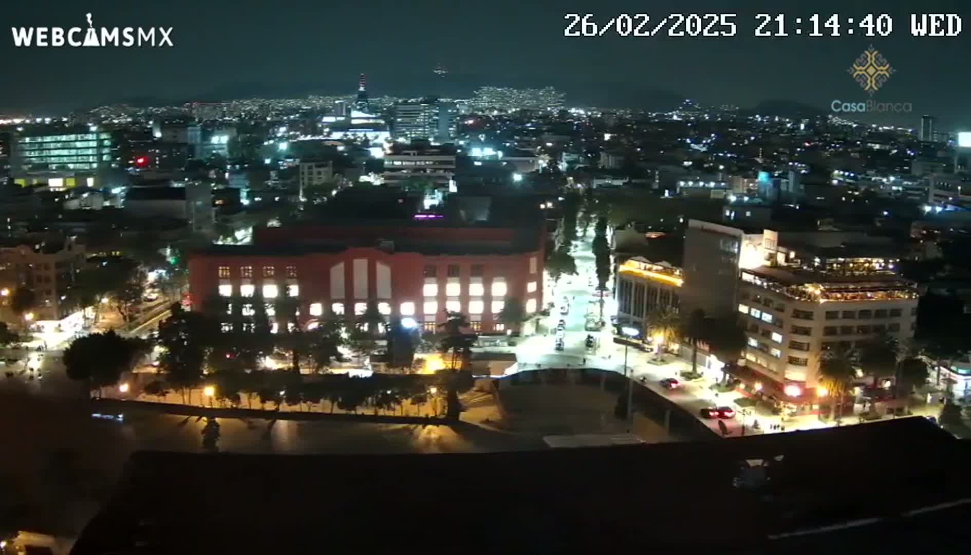 A nighttime aerial view of a city shows brightly lit buildings and streets, with trees and a large plaza visible below.