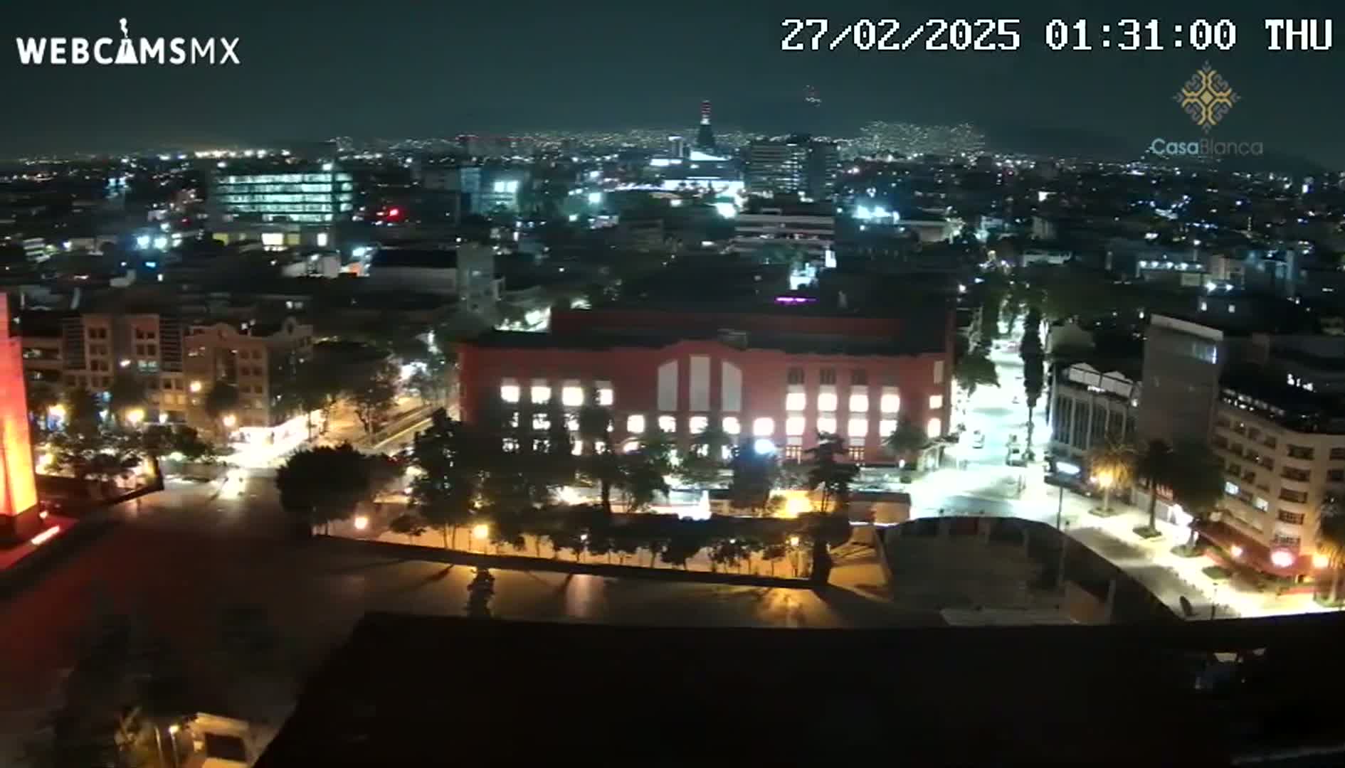 A nighttime, high-angle view of a city with brightly lit buildings and streets, showing a large square and a red building in the center.