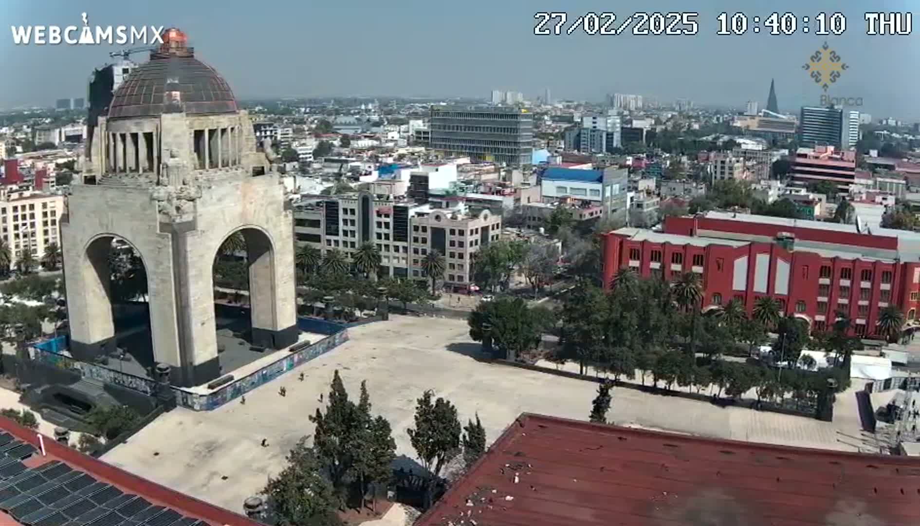 A large stone monument with arches stands in a city square on a clear day, surrounded by buildings and trees.
