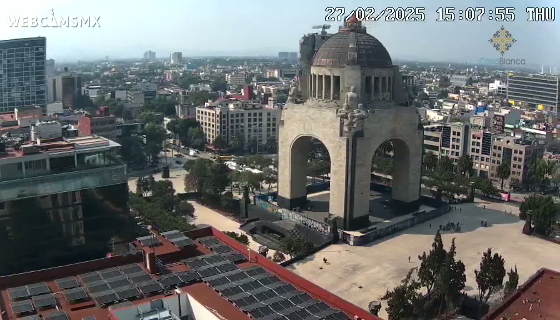A high-angle, sunny view of a large monument in a city square, surrounded by buildings and trees.