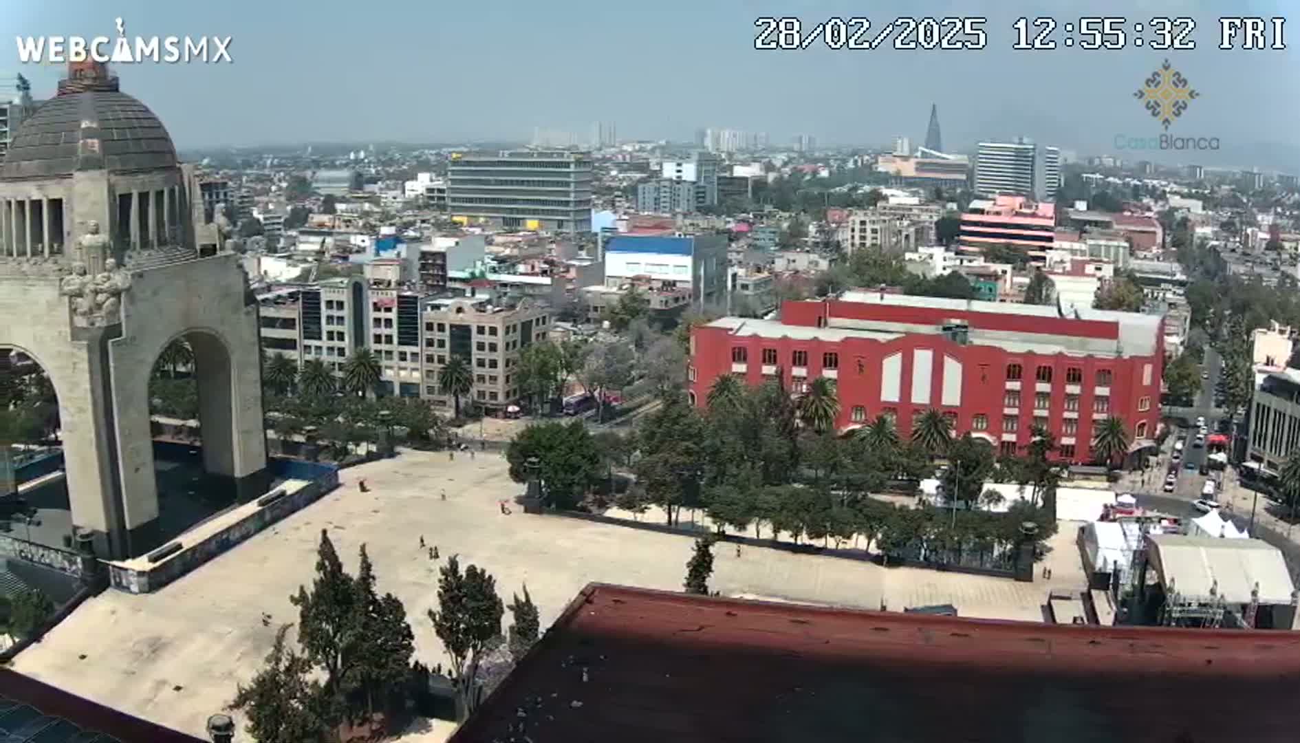 A high-angle view on a clear day shows a large plaza with a monument in the foreground, surrounded by a city with buildings of varying heights and a large red building.
