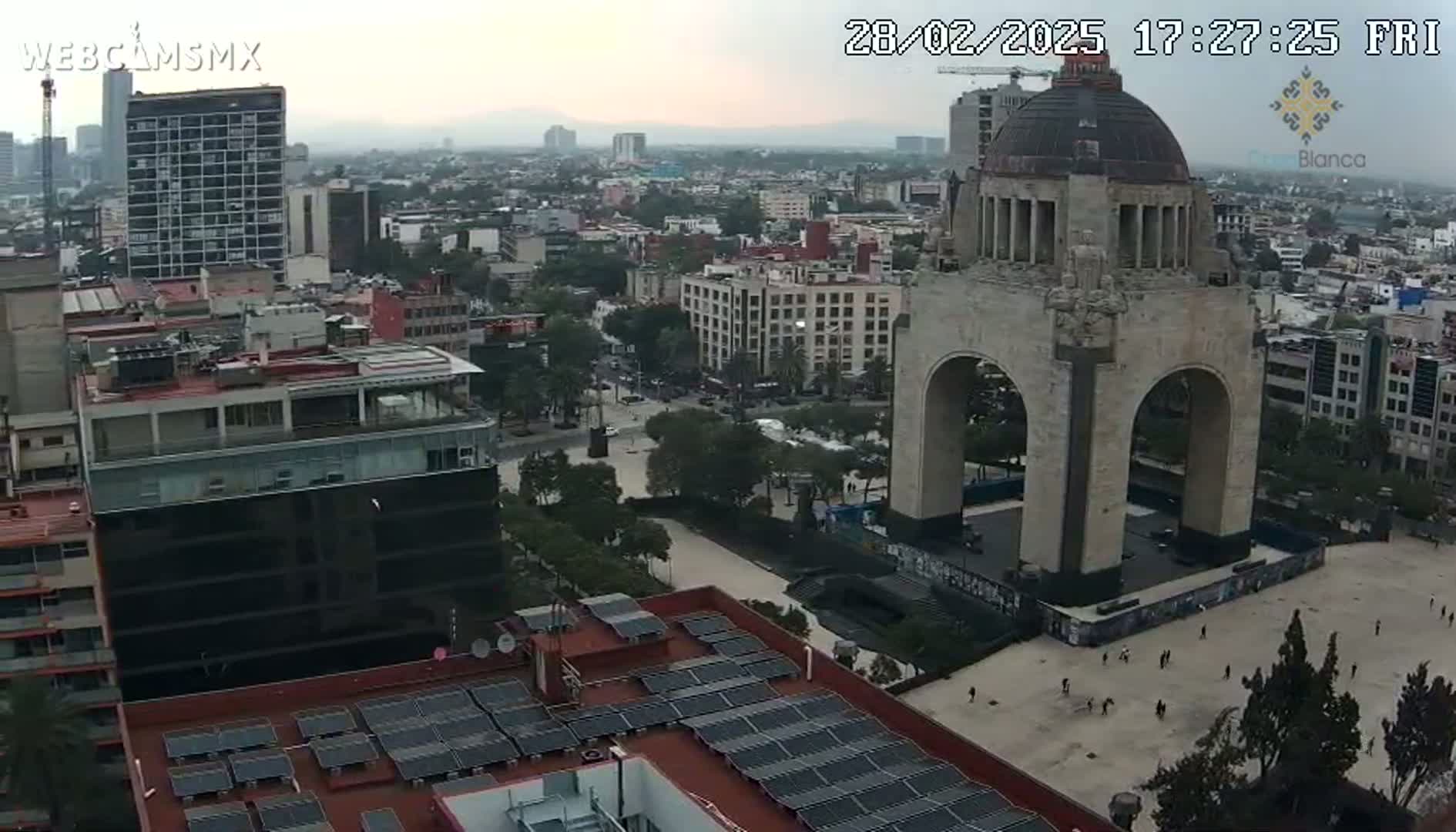 An elevated view of a city on a hazy day shows a large monument with two arches in a plaza, surrounded by buildings and a few scattered people.