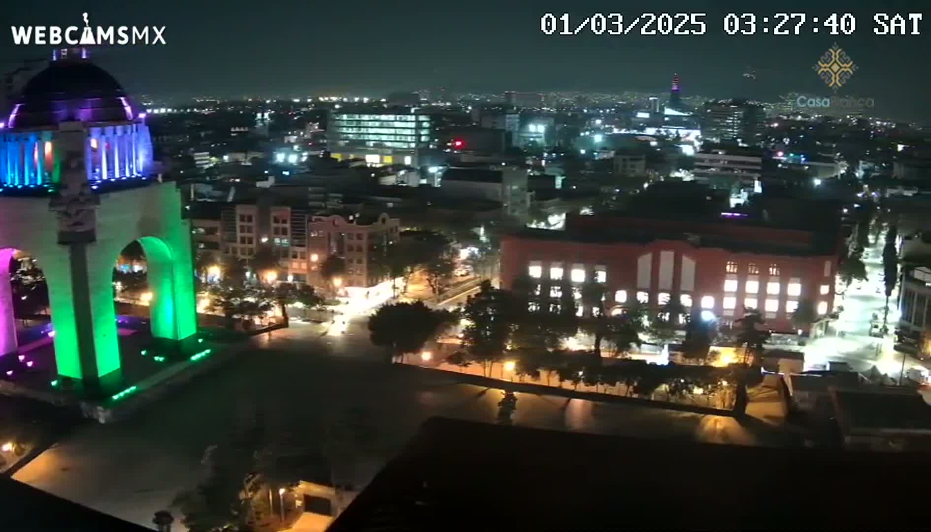 A nighttime, high-angle view of a city illuminated by streetlights, featuring a large monument with green and purple lighting in the foreground.