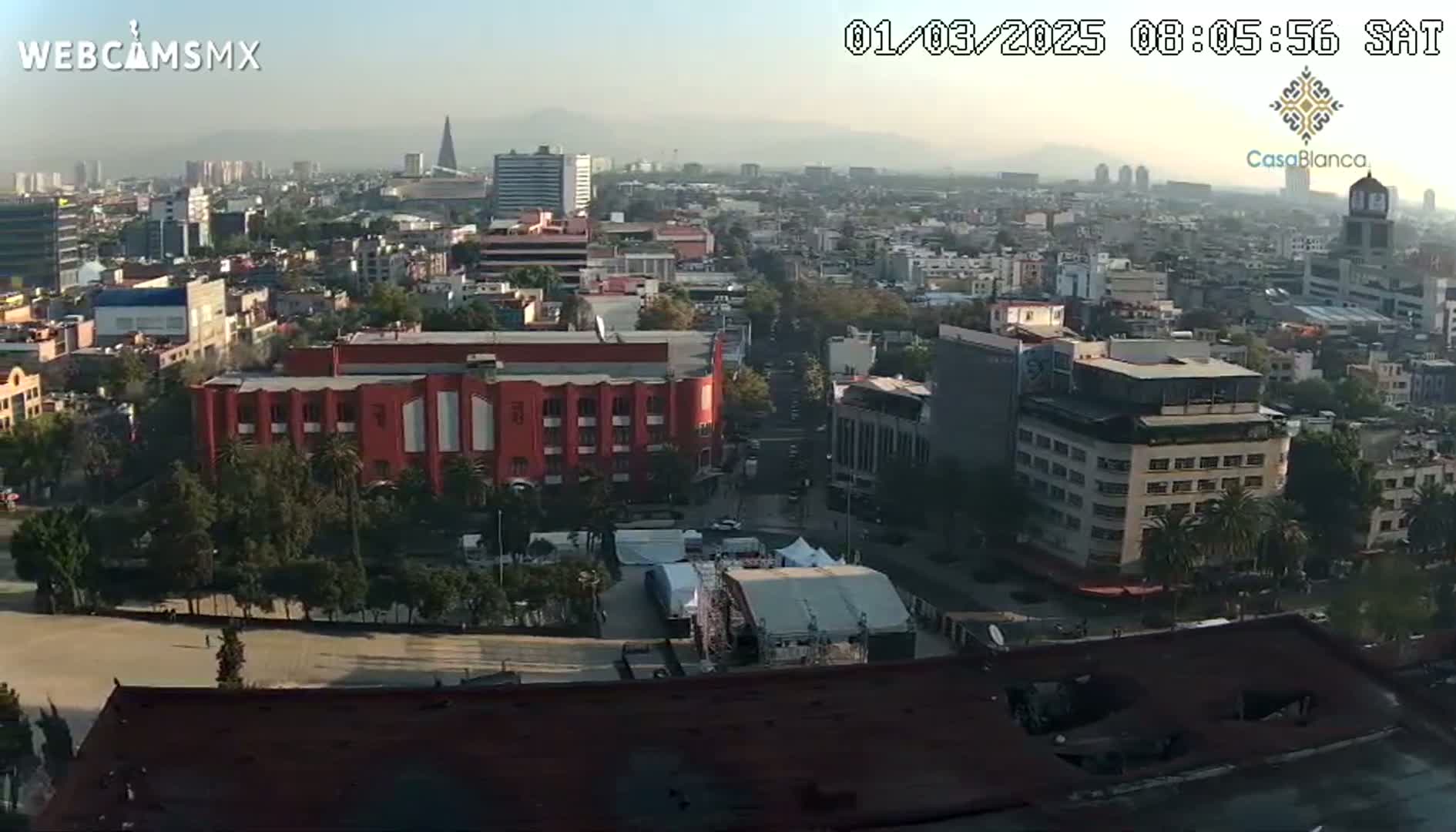 A hazy, sunny day view of a sprawling city, showing a mix of residential and commercial buildings, with a large red building prominent in the mid-ground and a stage setup in a plaza.