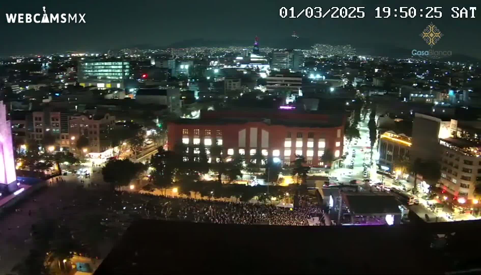 A nighttime aerial view shows a large crowd gathered in a city square, surrounded by illuminated buildings and streets.