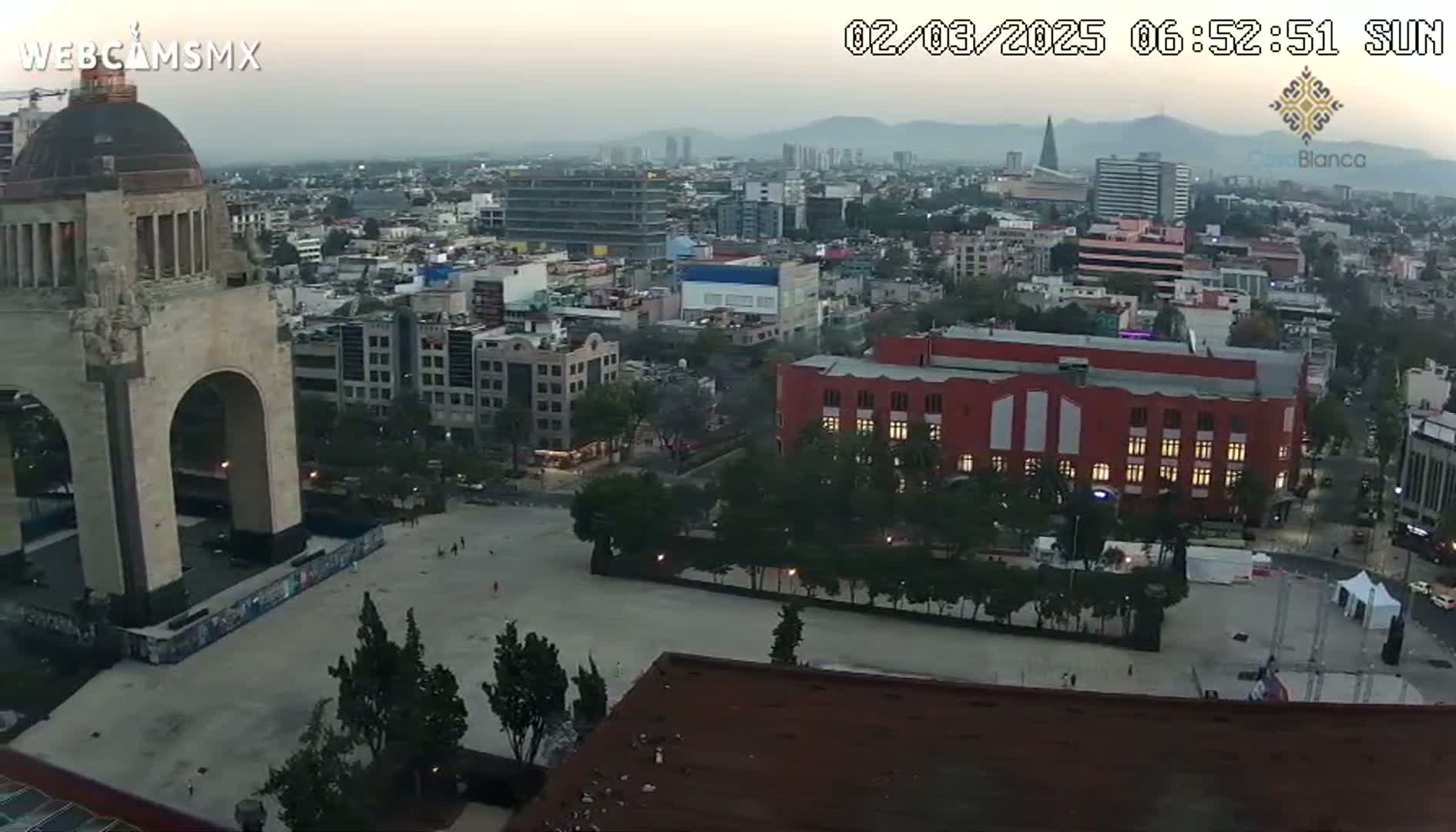 A high-angle, twilight view of a city square dominated by a large stone archway, with surrounding buildings and a hazy sky.