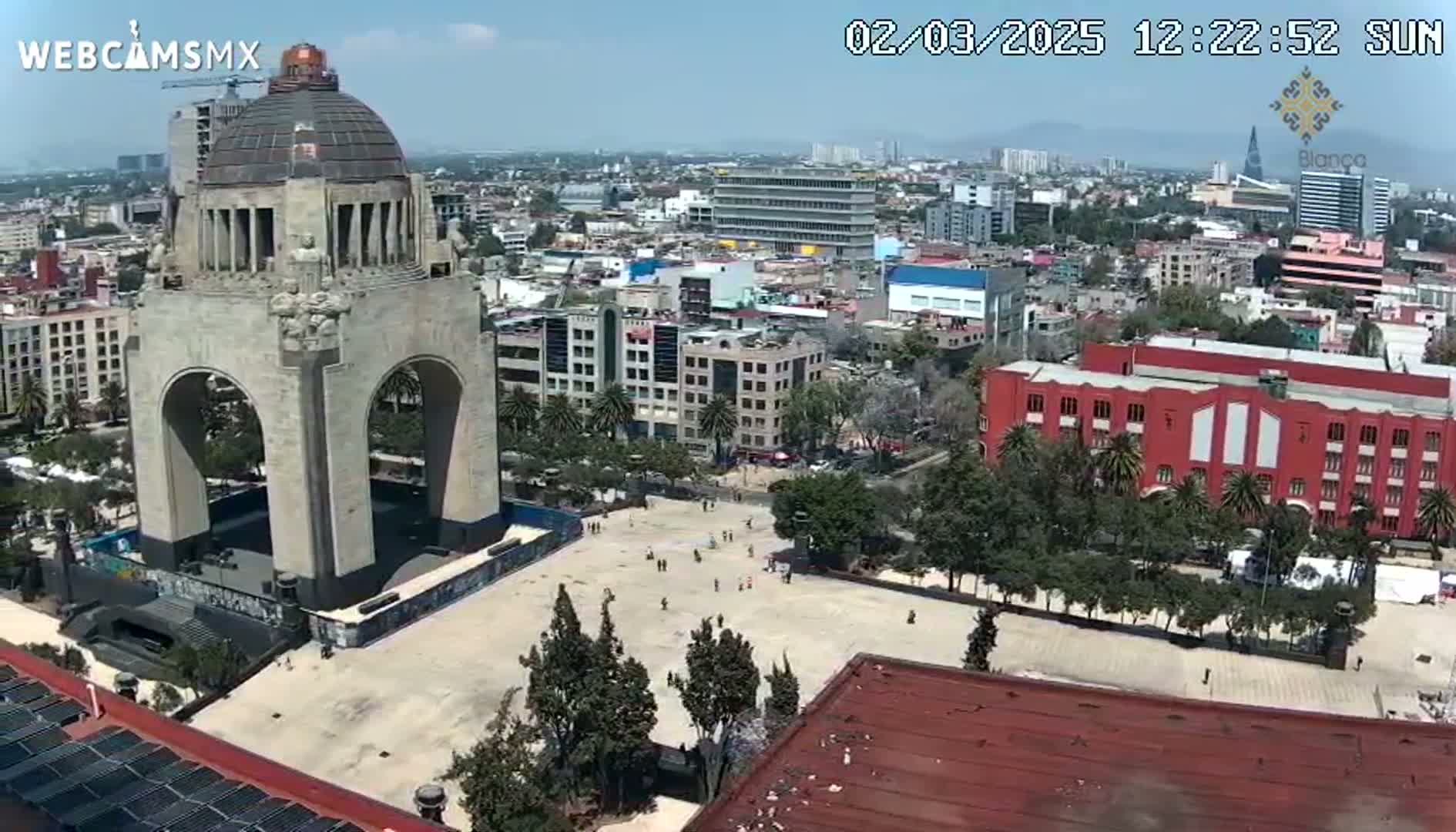 A large monument with an archway sits in a city square on a sunny day, surrounded by buildings and trees.