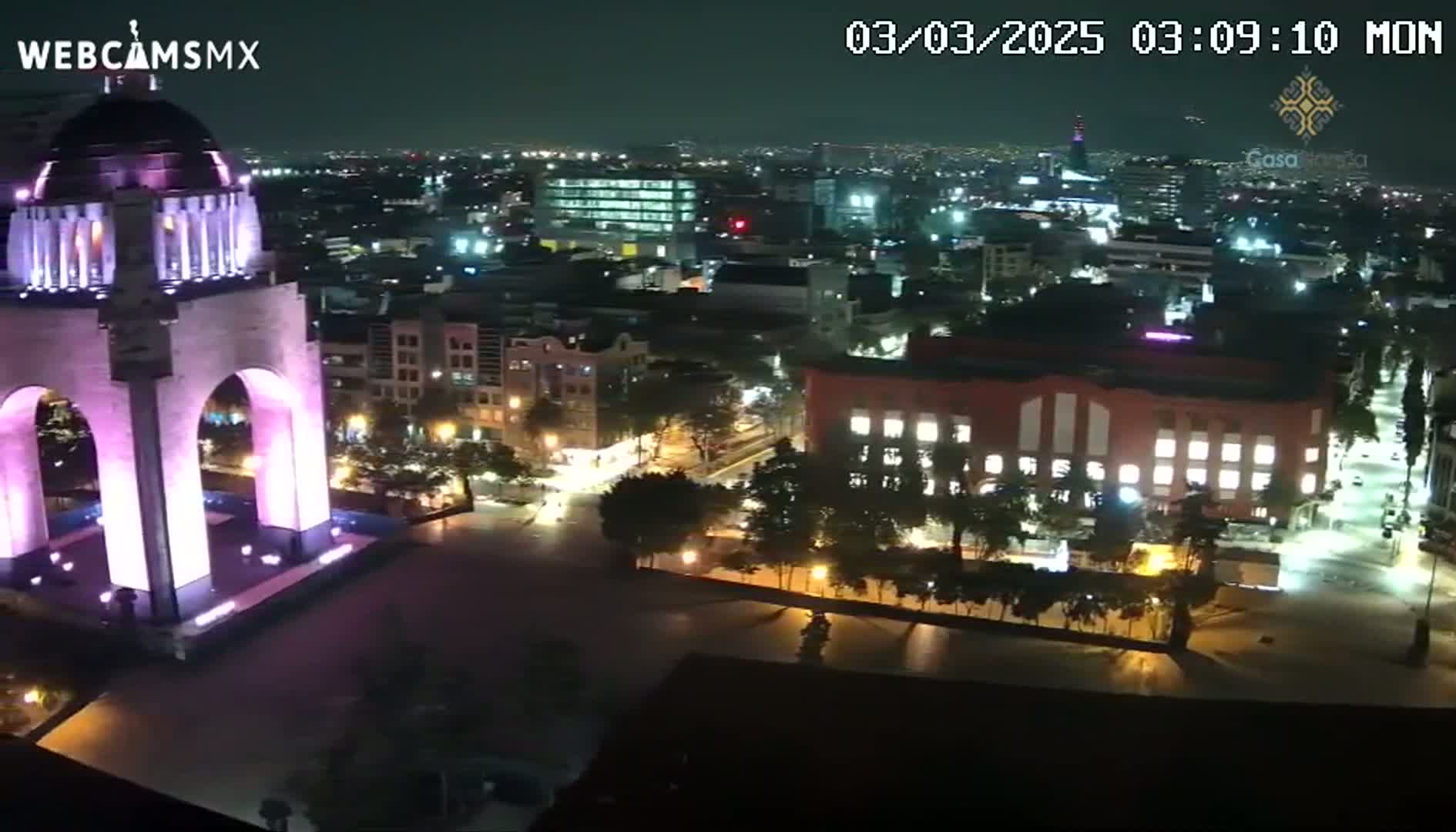 A nighttime, high-angle view of a city with a large monument illuminated in purple, surrounded by buildings and streets with sparse traffic.