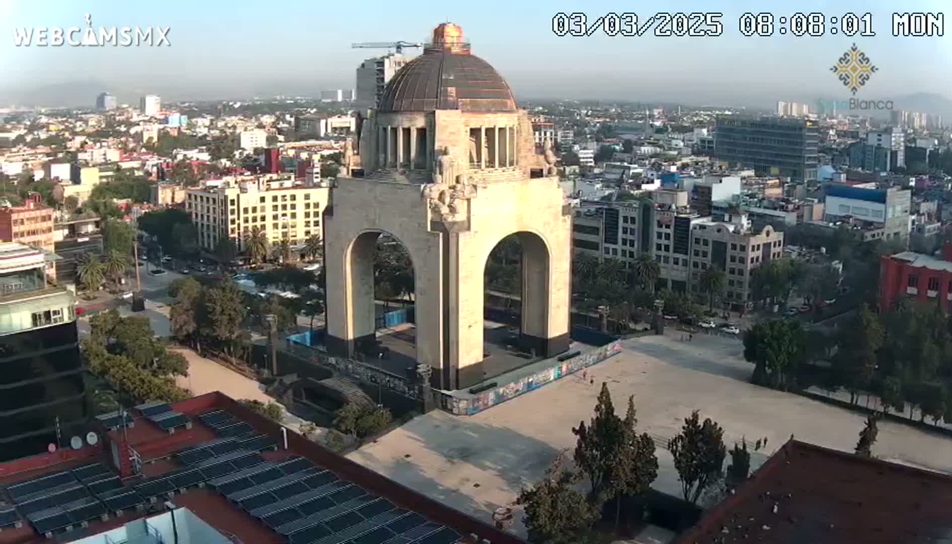A large, light-colored stone arch monument stands in a city square on a hazy, sunny day.
