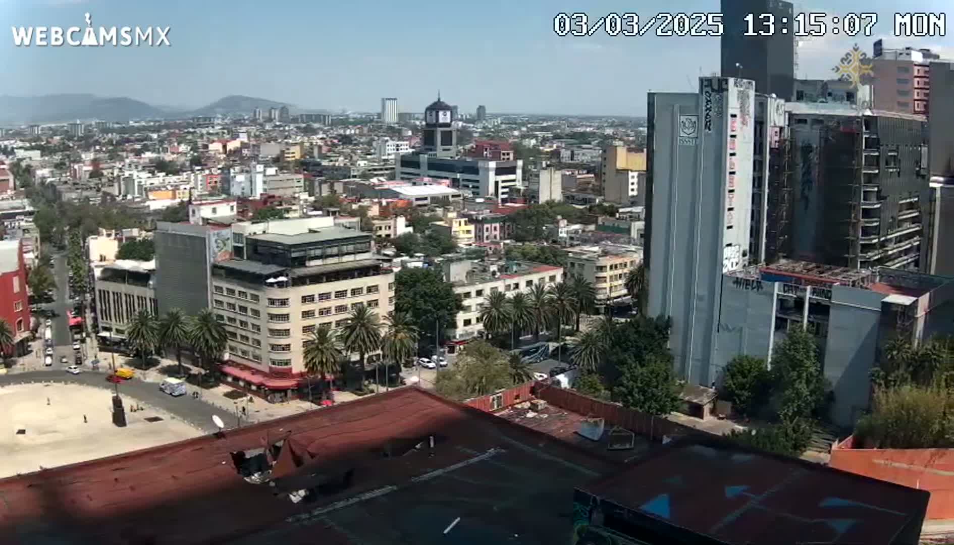 A sprawling cityscape under a clear, sunny sky, showing various buildings, streets, and palm trees.