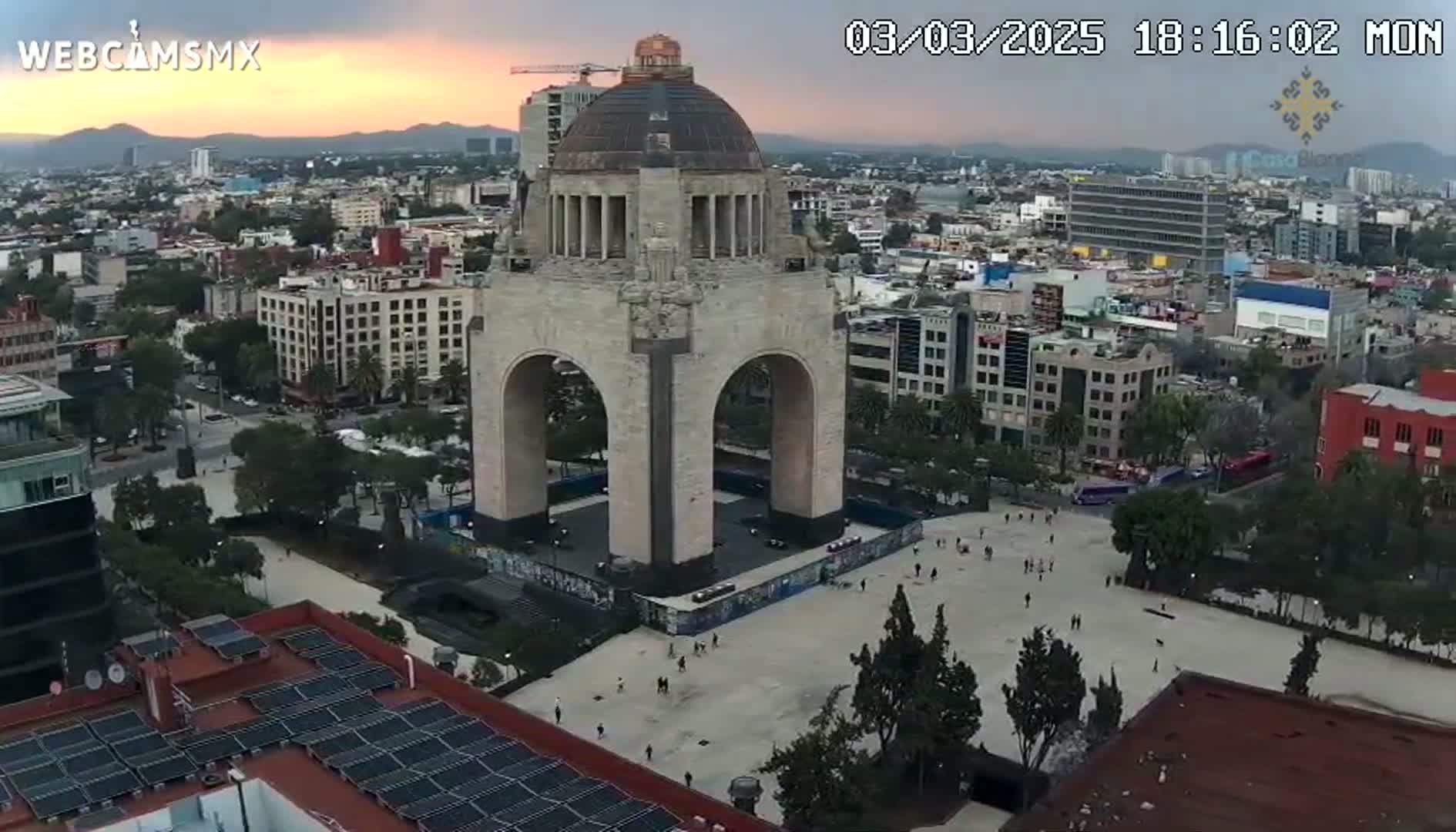 An aerial view shows a large monument with an archway in a city square, under a partly cloudy sunset sky.