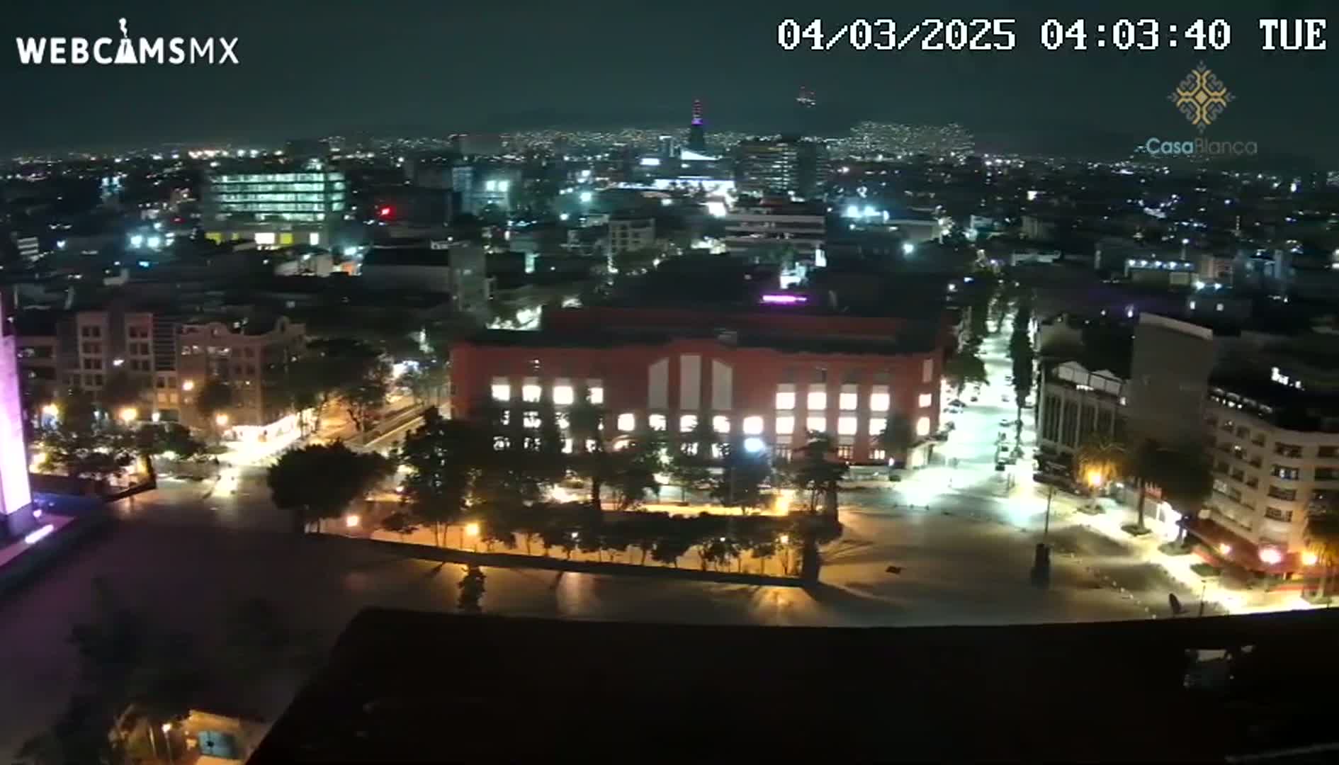 A nighttime aerial view of a city, showing brightly lit streets, buildings, and a large plaza.