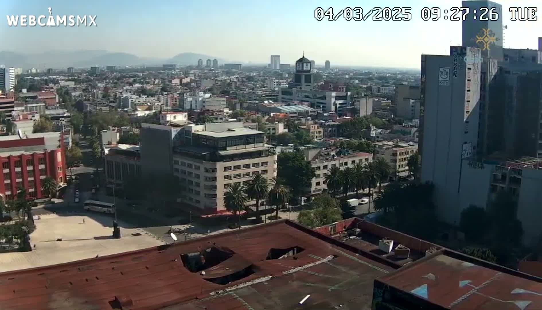 A high-angle, long shot on a sunny day shows a sprawling city with a mix of low and high-rise buildings, a large plaza, and palm trees, all under a clear sky.