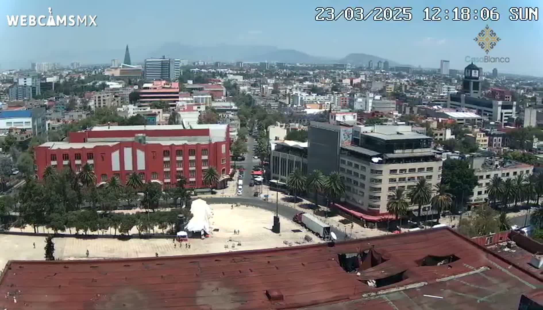 A sunny day reveals a wide shot of a city square and surrounding buildings, with palm trees lining the streets and a large red building prominently featured.