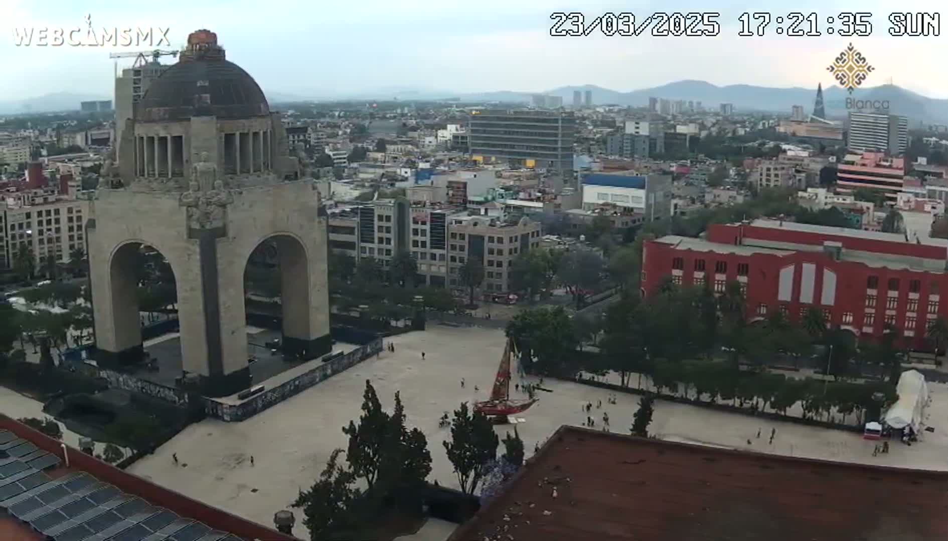 An elevated view of a large monument in a city square, under an overcast sky, with surrounding buildings and trees.
