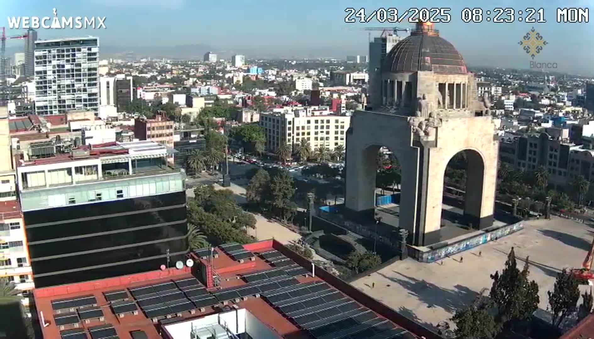 A high-angle, long shot on a clear day shows a large monument with an archway in a city with many buildings, some with solar panels on their roofs.