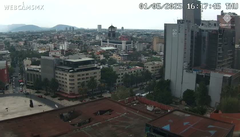 A high-angle, long shot of a city on an overcast day shows many buildings, streets, and a large square in the foreground.